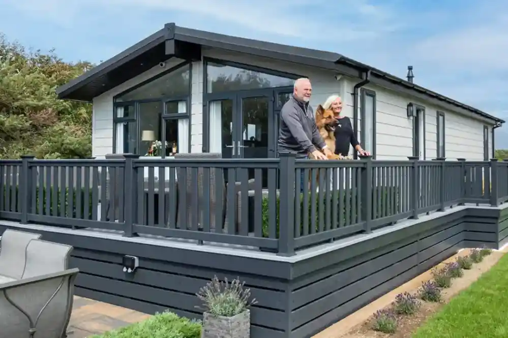 A couple stands on the deck of a modern, gray-hued vacation home, smiling as they hold a small dog. The home features large windows and a manicured landscape in the background, with a blue sky above.