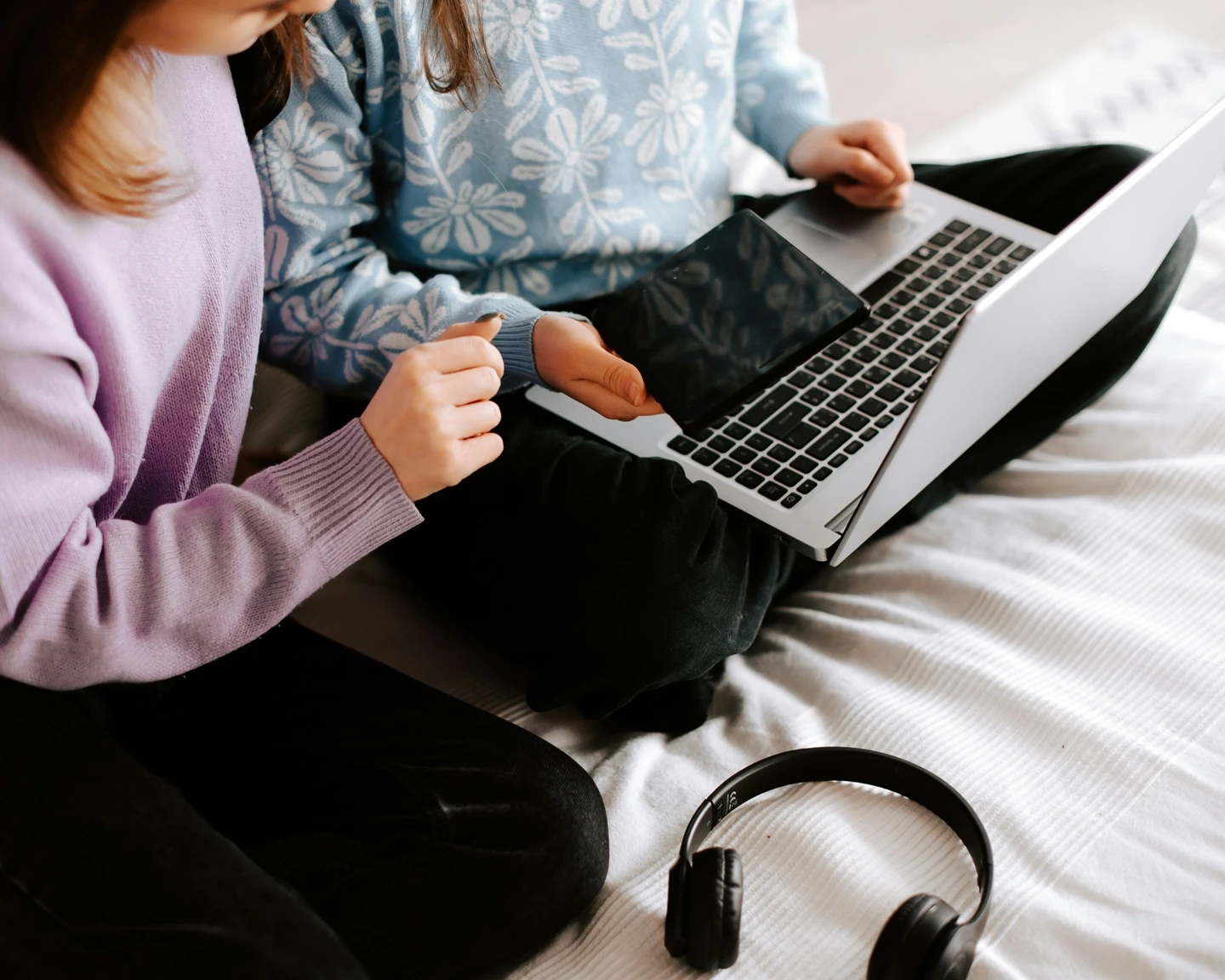 Two kids on a laptop sat on a comfy bed. There is a set of headphones in the foreground.